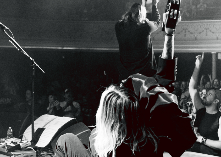 Black and white image of the members of 'Band of Skulls' on stage performing taking from behind them and the crowd is in front with a man in the bottom right with his hand raised in the air.x